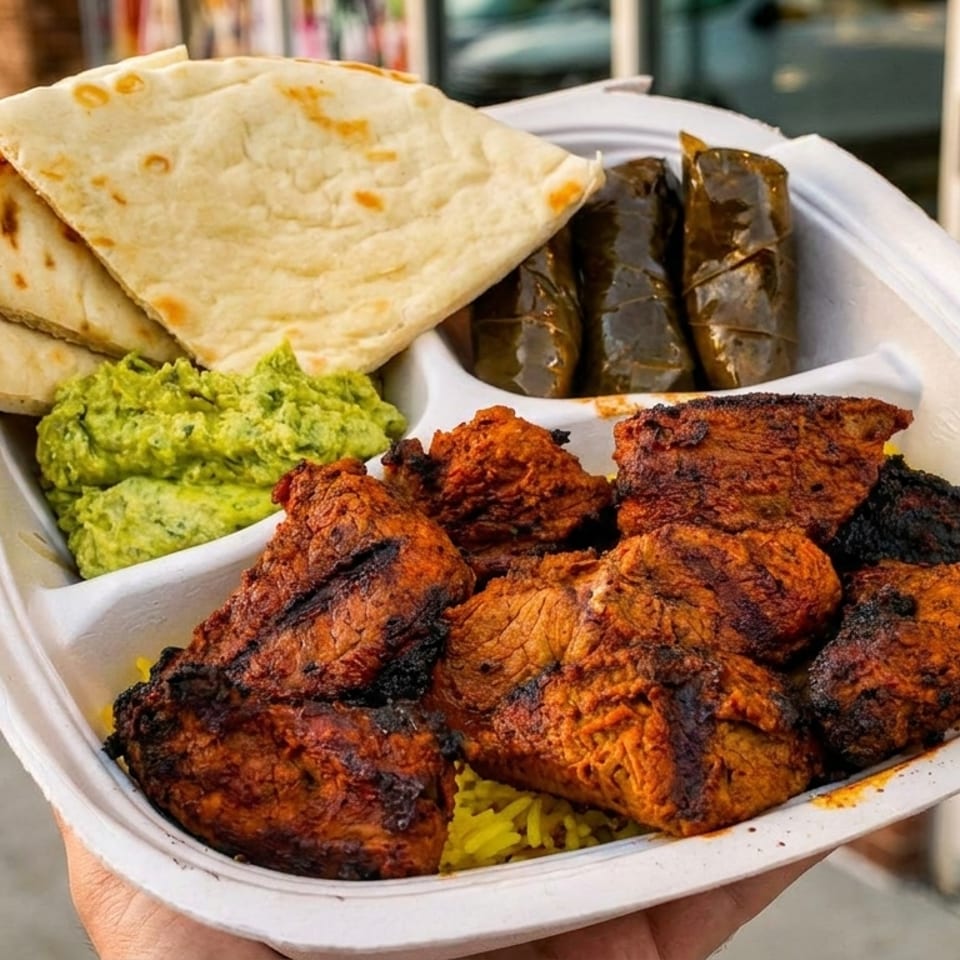 Takeout platter featuring grilled spiced chicken, yellow rice, flatbread, green dip, and stuffed grape leaves.