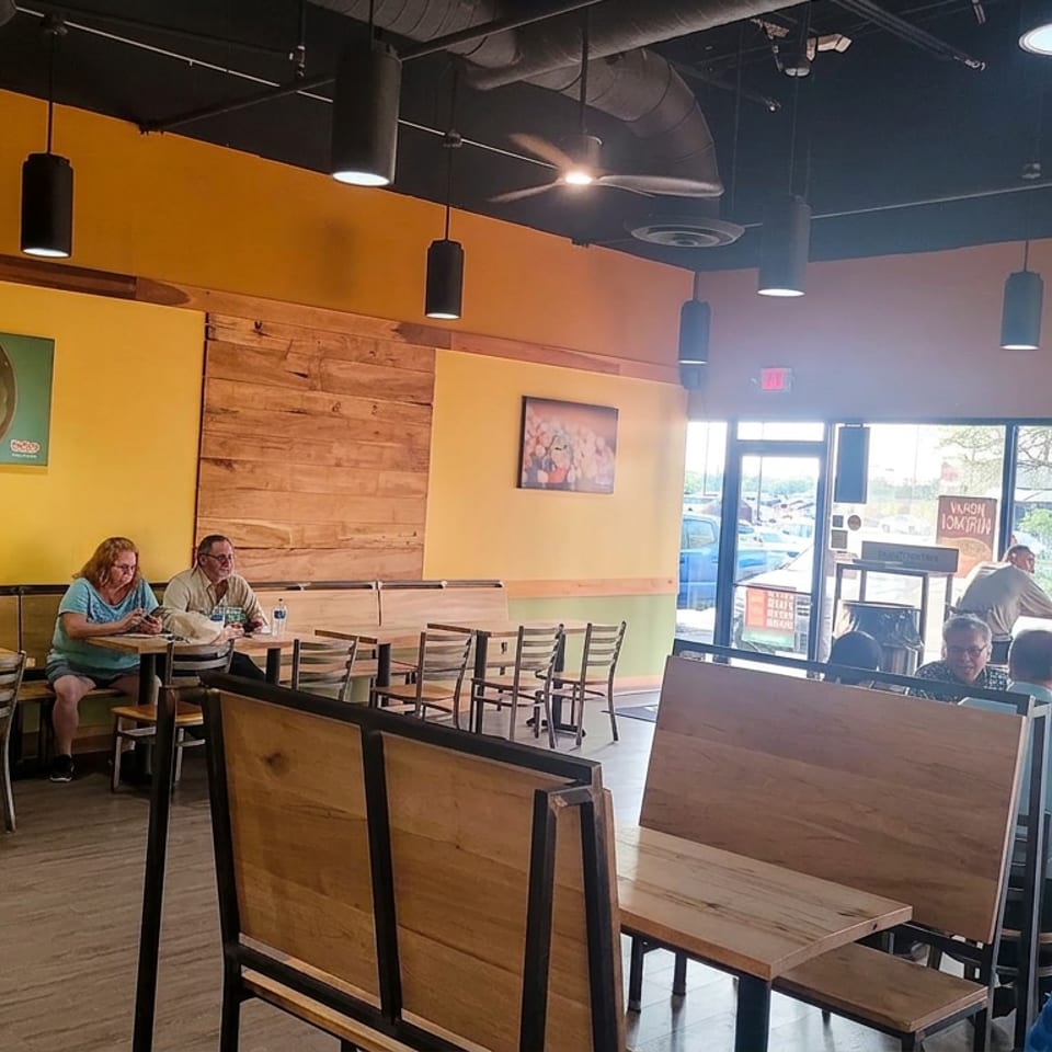 Bright casual restaurant dining room with wooden tables, colorful walls, pendant lights, and customers seated near large windows.