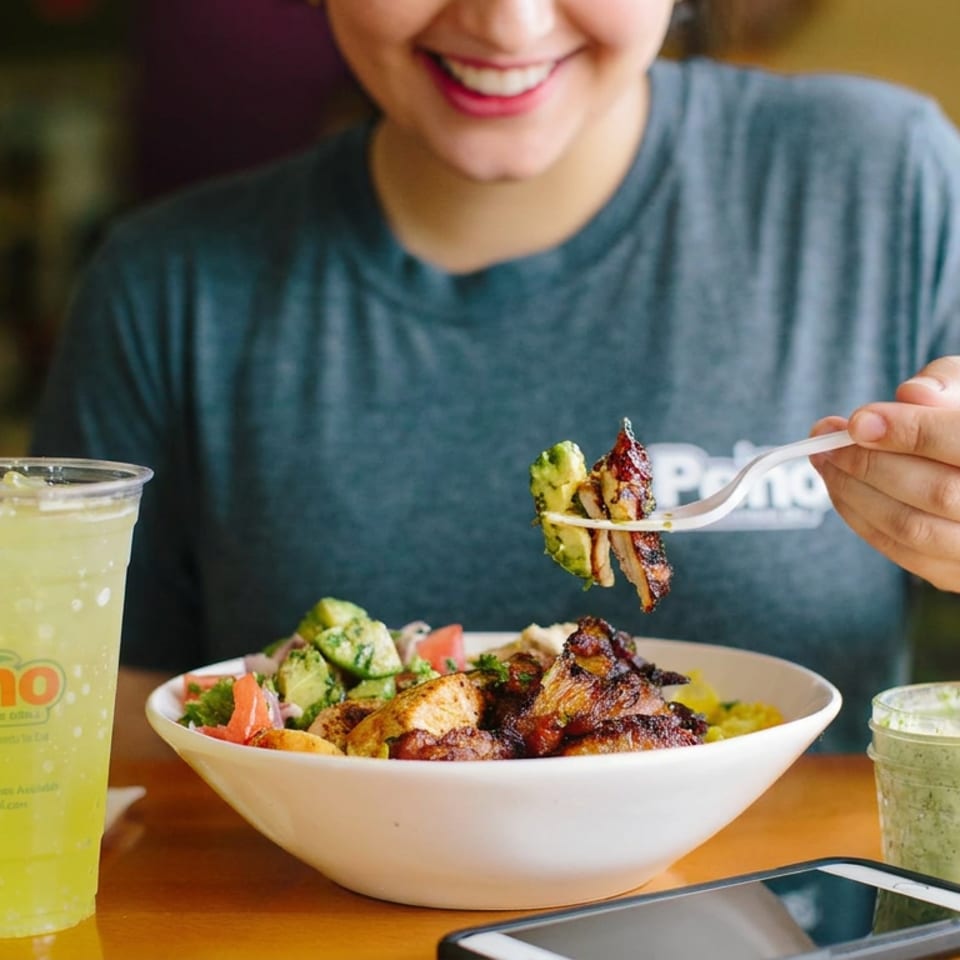 Diner lifts grilled meat and vegetables from a bowl beside a Peno drink, green sauce, and smartphone.