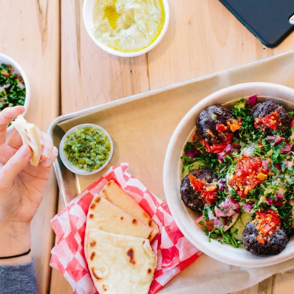Falafel bowl topped with salad and roasted vegetables, served with pita, dipping sauce, and a hand-held piece of bread.