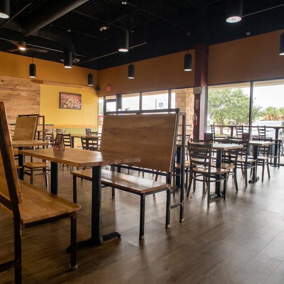 Bright casual restaurant dining area with wooden tables, black chairs, colorful food artwork, and floor-to-ceiling windows.