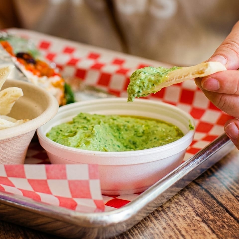 Hand dips pita bread into green hummus on a red-and-white checkered restaurant tray.