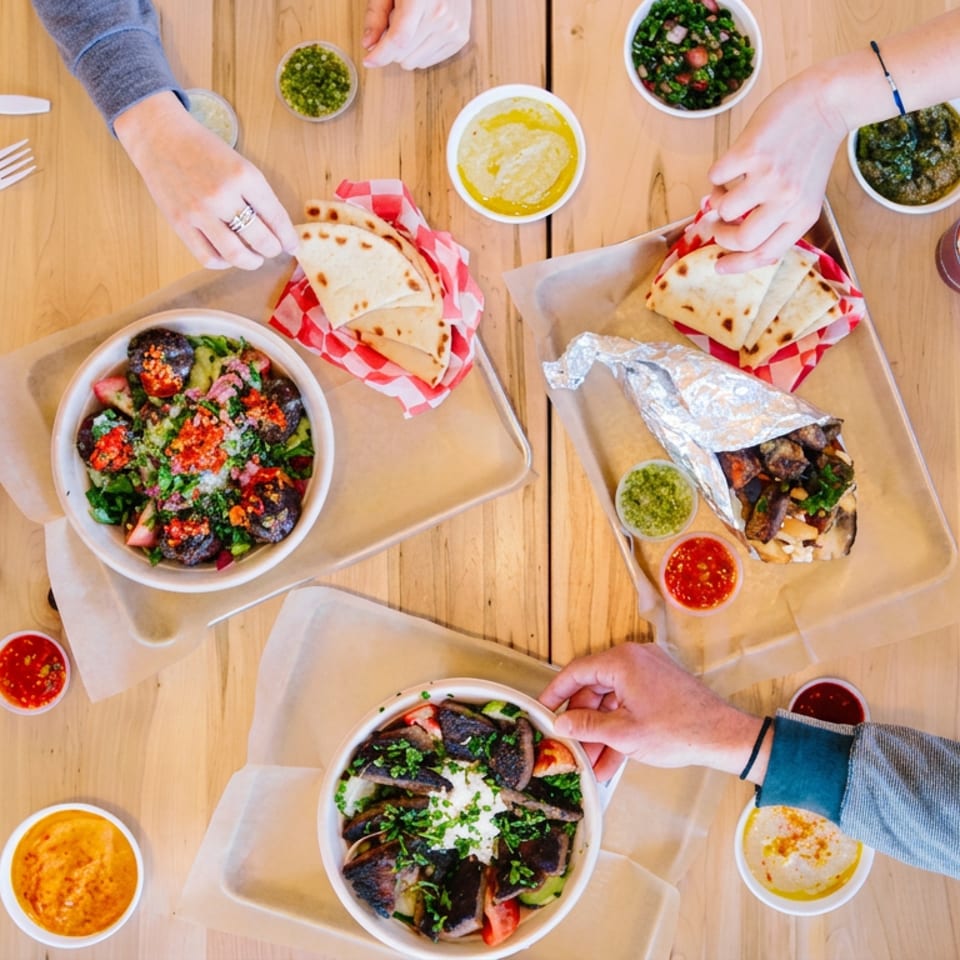 Overhead view of diners sharing Mediterranean dishes, flatbreads, dips, salads, sauces, and drinks on a wooden table.