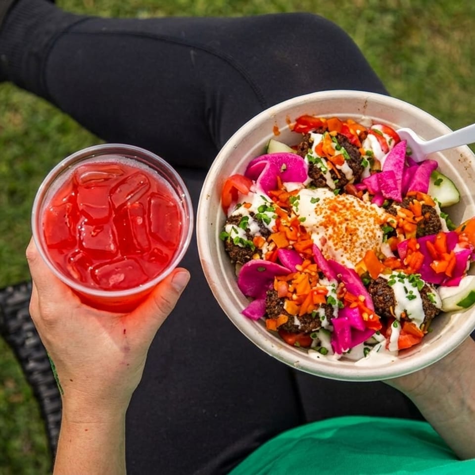 Person seated outdoors holds a colorful falafel bowl with hummus, pickled vegetables, and a red iced drink.