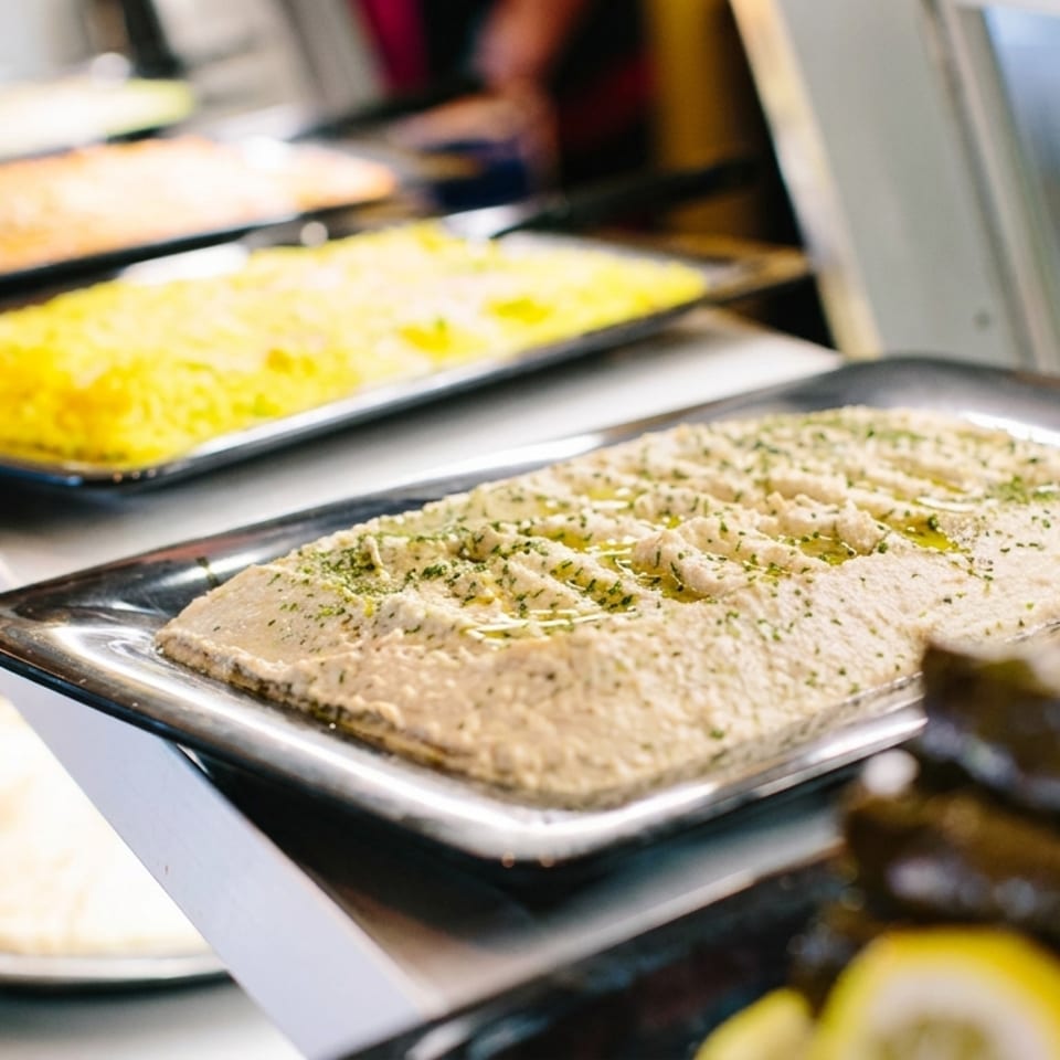 Buffet display featuring herb-topped hummus, yellow rice, roasted vegetables, and assorted dishes in stainless steel trays.