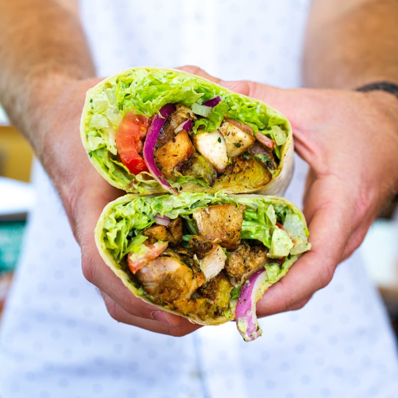 Hands holding a Peno Mediterranean Grill wrap, cut in half, filled with chicken, lettuce, and vegetables.