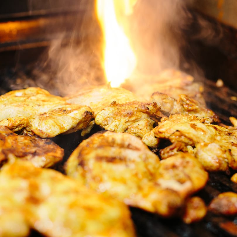 Chicken pieces grilling on a hot griddle with flames and smoke rising from the heat.