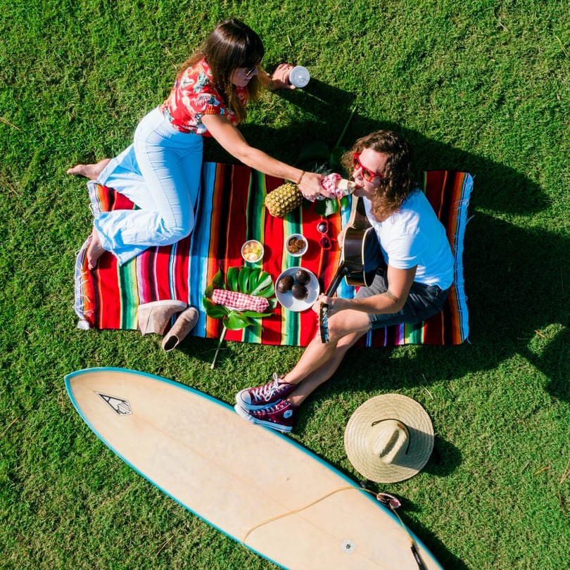 Overhead view of two people picnicking on a colorful blanket with fruit, snacks, and a surfboard nearby.